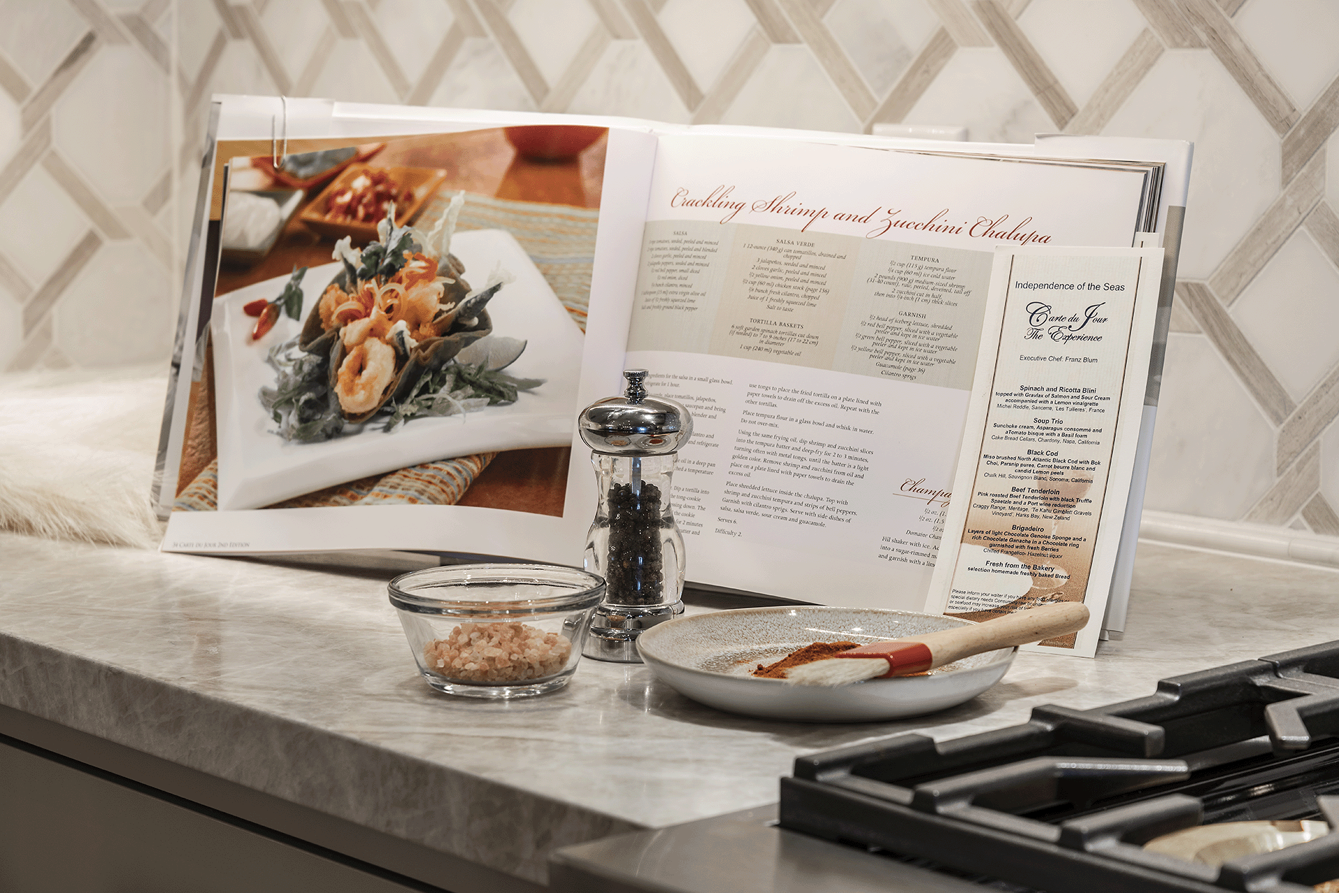 Cookbook open on kitchen counter next to stove, with a bowl of salt and pepper grinder.