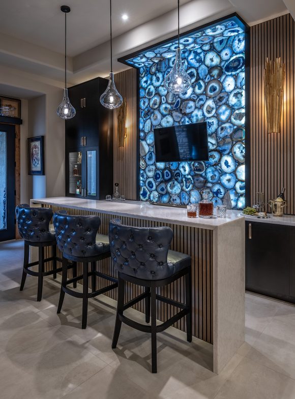 Bar with backlit wall design, mounted T.V., three leather barstools at island and hanging lights.